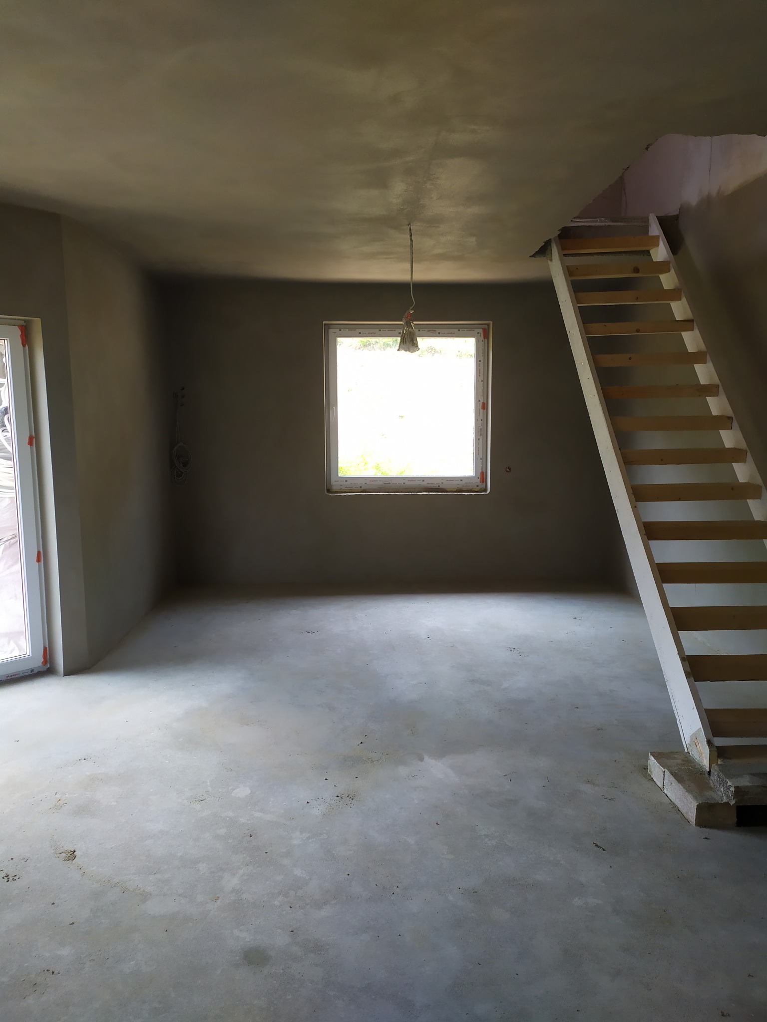 Surowe wnętrze domu w trakcie prac wykończeniowych: ściany pokryte świeżym tynkiem, drewniane schody bez poręczy, surowa podłoga i wisząca żarówka.
