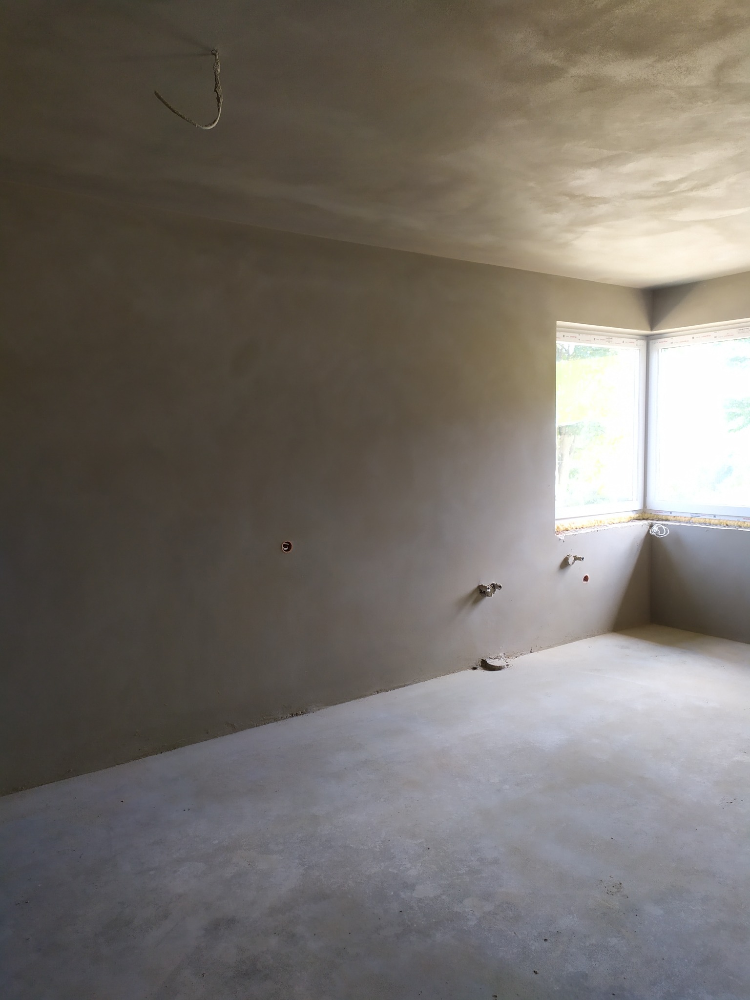 Surowe wnętrze pokoju po tynkowaniu maszynowym, widoczne przygotowane instalacje elektryczne i hydrauliczne, narożne okno z widokiem na zieleń.