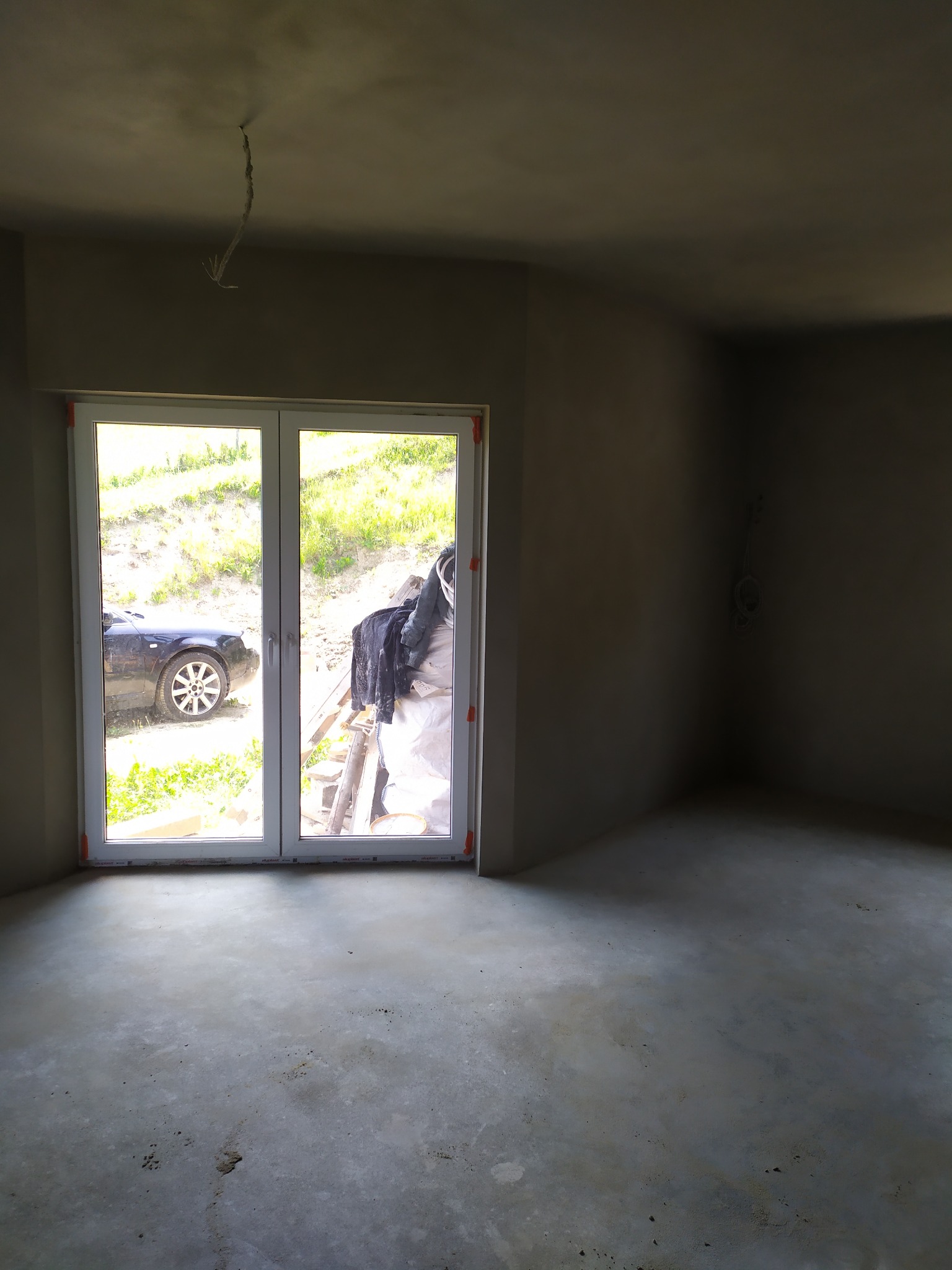 Surowe wnętrze po nałożeniu tynku, widoczne kable wystające z sufitu, okno z widokiem na samochód i teren zielony.