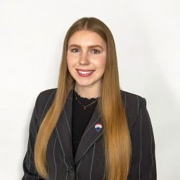 RE MAX Deluxe - Portret uśmiechniętej kobiety w ciemnym, prążkowanym żakiecie z przypinką agencji nieruchomości, na białym tle.