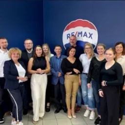 Grupa uśmiechniętych agentów nieruchomości pozuje na tle niebieskiej ściany z logo RE/MAX; widoczny podział kolorystyczny logo: czerwony, biały i niebieski.