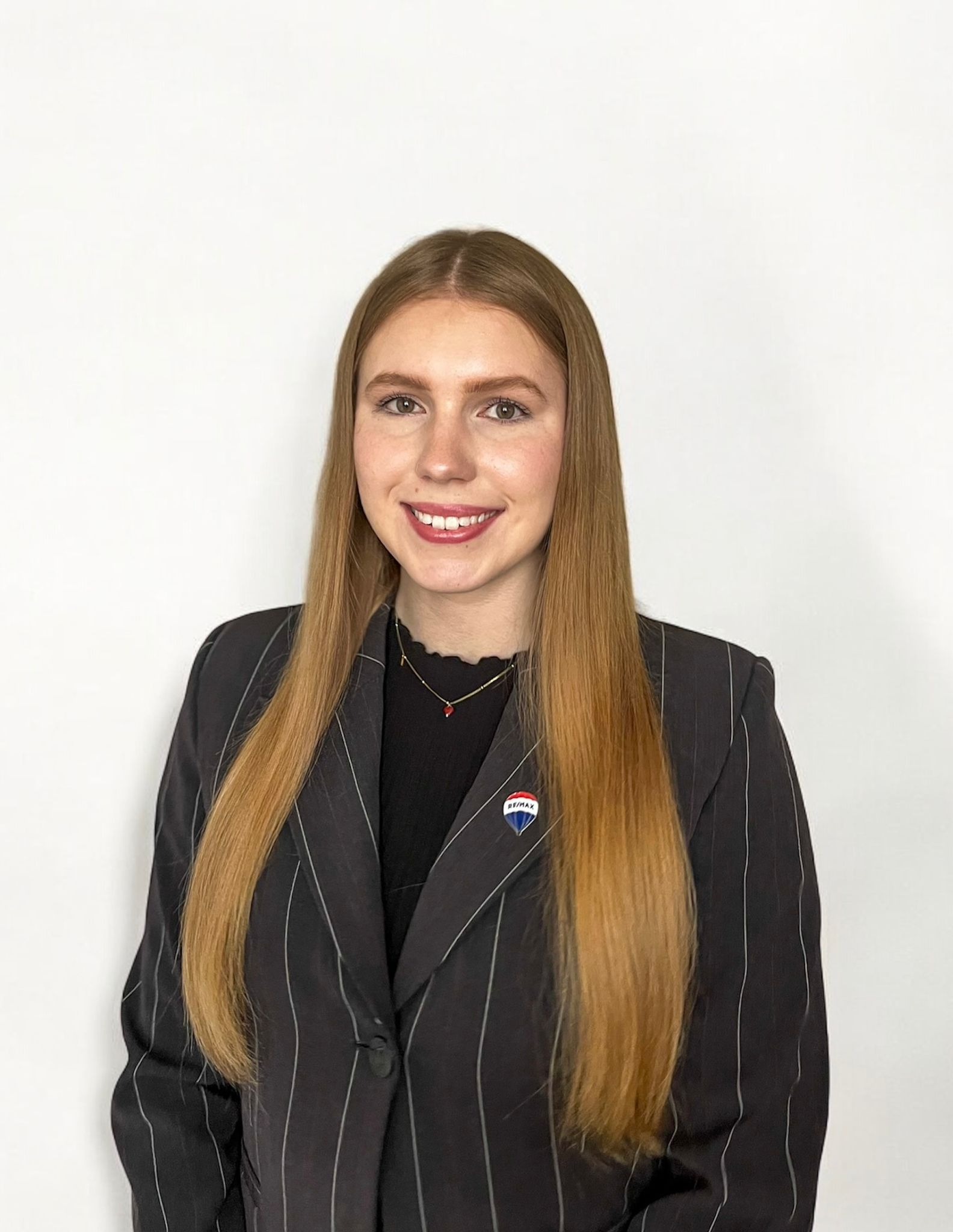Portret uśmiechniętej kobiety w ciemnym, prążkowanym żakiecie z przypinką agencji nieruchomości, na białym tle.