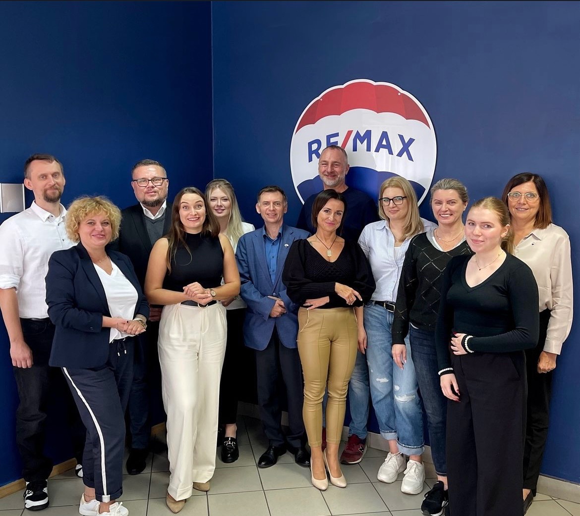 Grupa uśmiechniętych agentów nieruchomości pozuje na tle niebieskiej ściany z logo RE/MAX; widoczny podział kolorystyczny logo: czerwony, biały i niebieski.