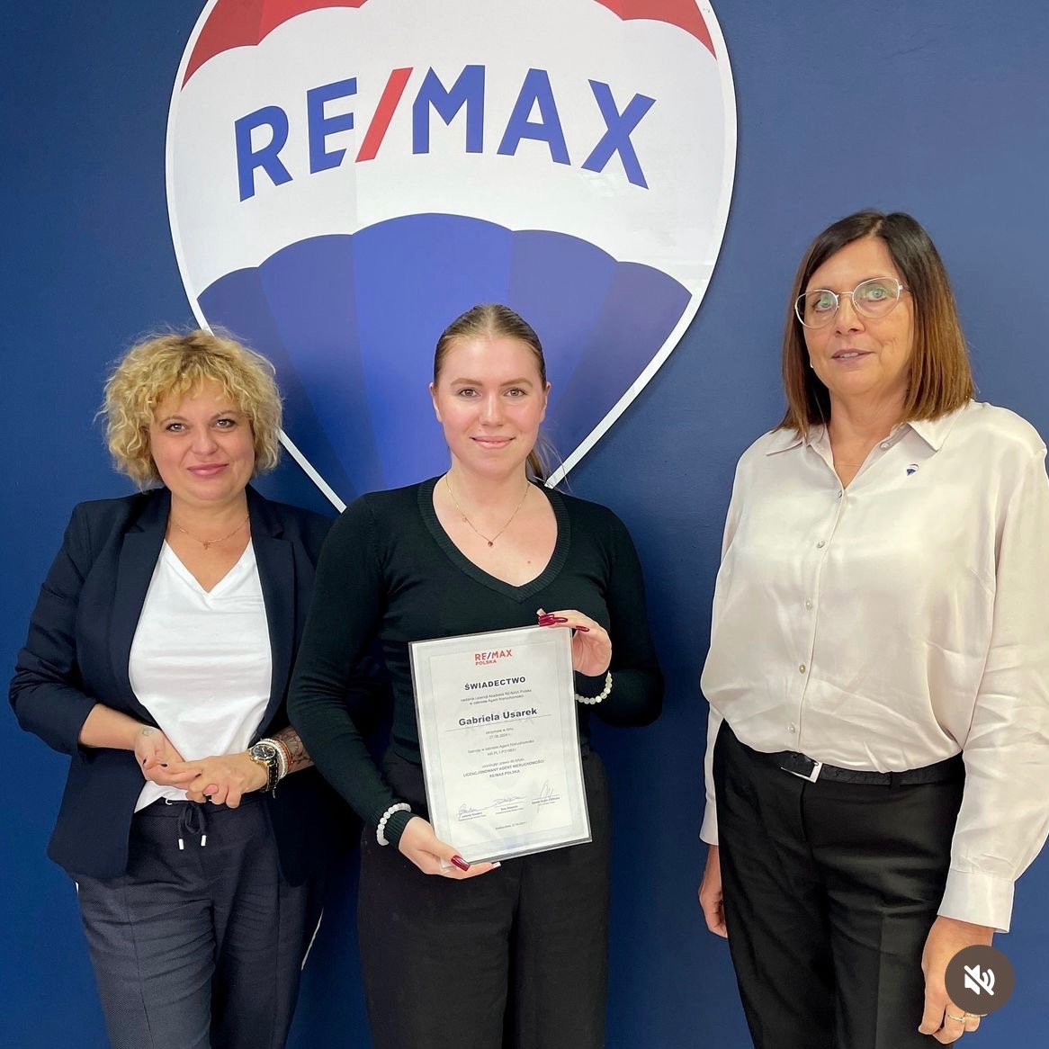 Trzy kobiety pozują na tle ściany z logo RE/MAX, środkowa trzyma certyfikat z imieniem Gabriela Usarek.