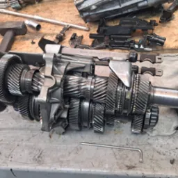 Rozłożona skrzynia biegów samochodowej na brudnym stole warsztatowym, widoczne narzędzia i elementy skrzyni biegów.