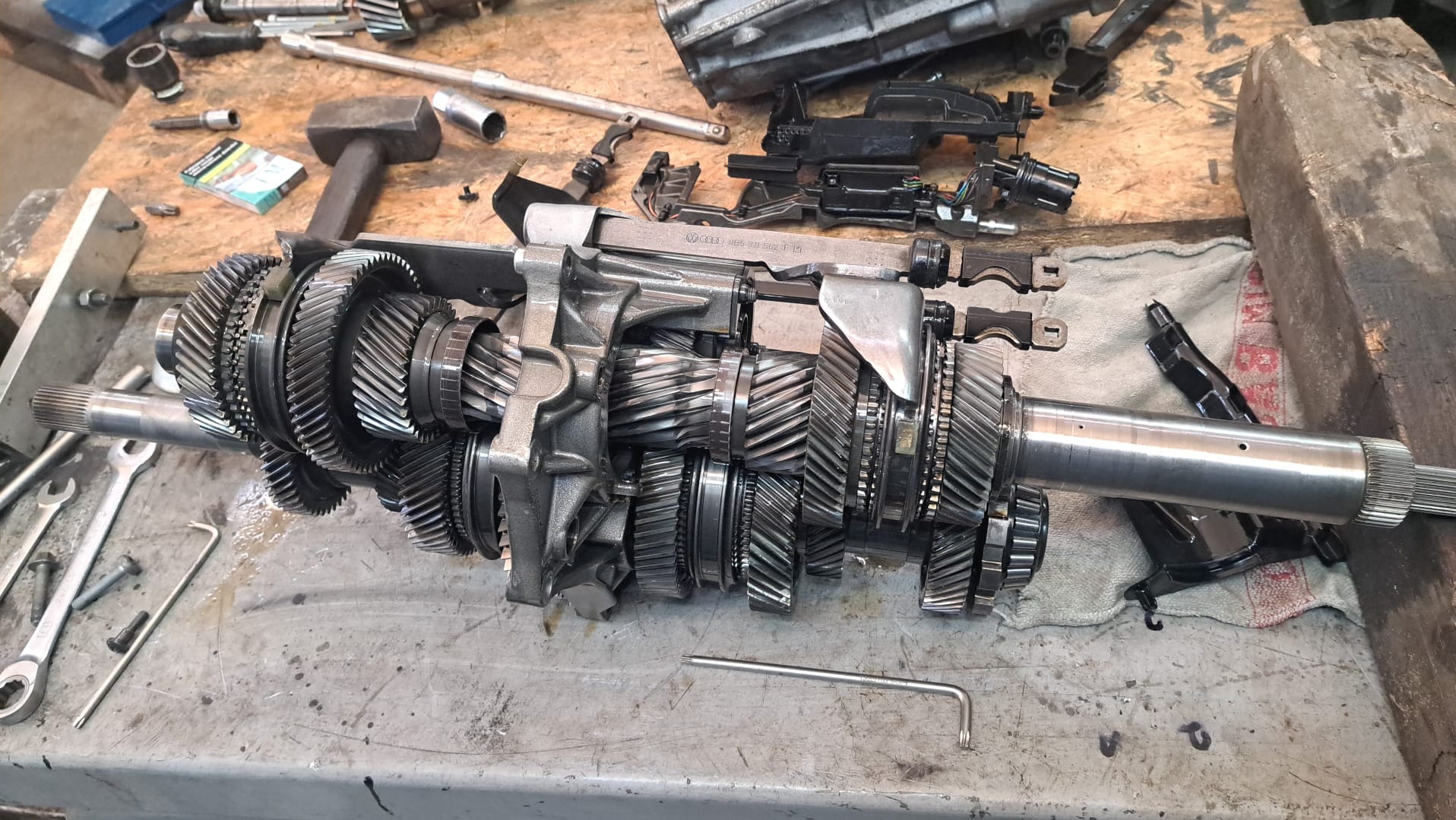 Rozłożona skrzynia biegów samochodowej na brudnym stole warsztatowym, widoczne narzędzia i elementy skrzyni biegów.