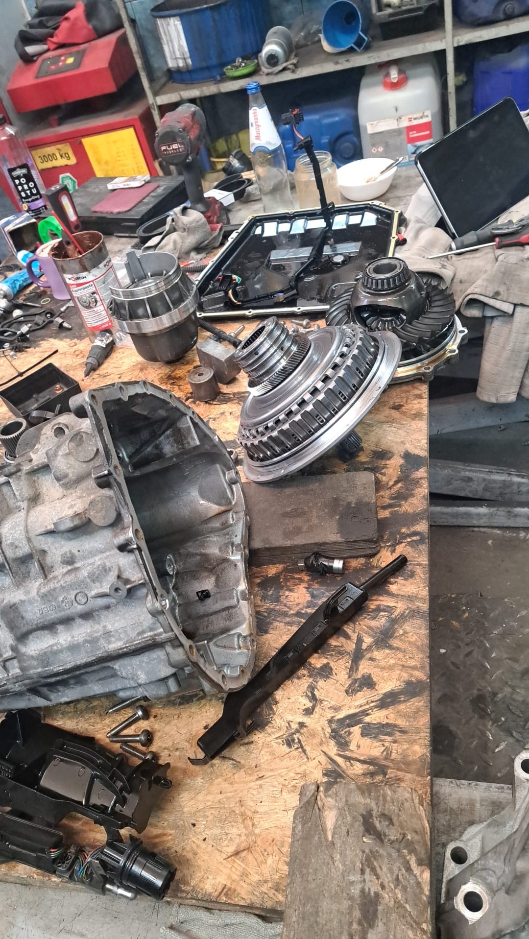 Rozłożona skrzynia biegów automatyczna na roboczym stole w warsztacie, widoczne elementy przekładni, obudowa, sterownik i narzędzia.