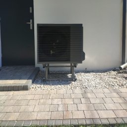 Ice Term - Czarna pompa ciepła Panasonic zamontowana na metalowej konstrukcji na zewnątrz budynku, obok ciemnych drzwi wejściowych i białej ściany, otoczona białymi kamieniami.