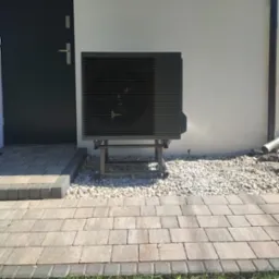 Czarna pompa ciepła Panasonic zamontowana na metalowej konstrukcji na zewnątrz budynku, obok ciemnych drzwi wejściowych i białej ściany, otoczona białymi kamieniami.