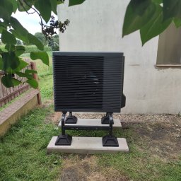 Ice Term - Czarna pompa ciepła Panasonic zamontowana na betonowych podstawach w trawie przed budynkiem z widocznym oknem i fragmentem drewnianego płotu.