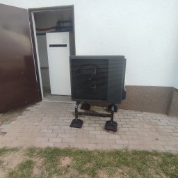 Ice Term - Czarna zewnętrzna jednostka pompy ciepła Panasonic Aquarea na metalowym stelażu, ustawiona na kostce brukowej przed białą ścianą budynku, z otwartymi drzwiami do pomieszczenia z wewnętrzną jednostką.