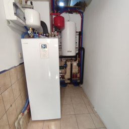 Ice Term - Biała pompa ciepła Nibe z podłączonymi rurami instalacji grzewczej, obok zbiornik Ferroli, widoczne elementy instalacji w ciasnym pomieszczeniu.