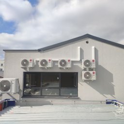 Ice Term - Biała ściana budynku z kilkoma zamontowanymi zewnętrznymi jednostkami klimatyzacji LG Dual Inverter, widoczne przewody instalacji i okno, ujęcie z góry z dachu innego budynku.