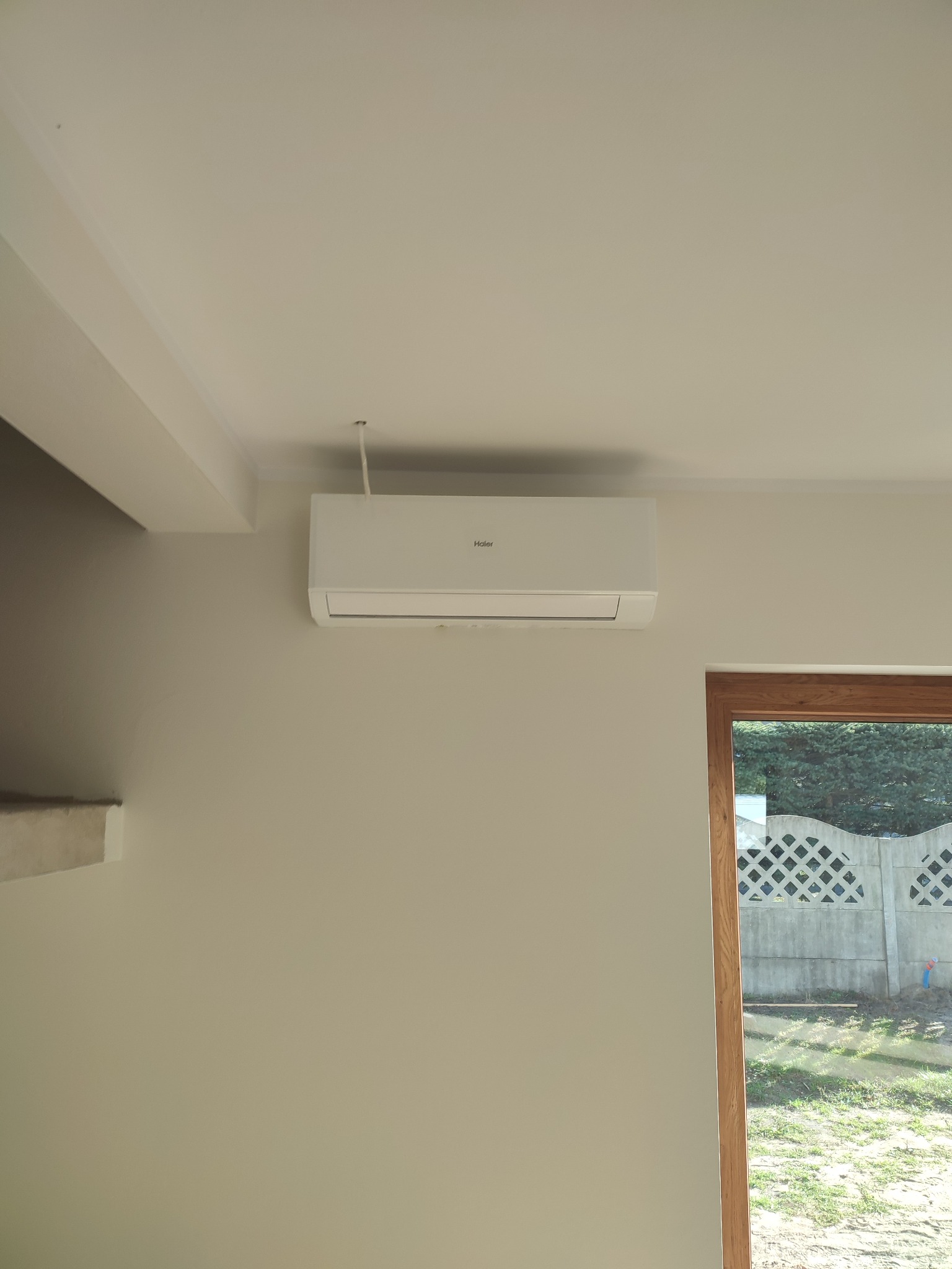 Biały klimatyzator Haier zamontowany na ścianie w jasnym pomieszczeniu, widoczne okno z drewnianą ramą i betonowe ogrodzenie za nim.