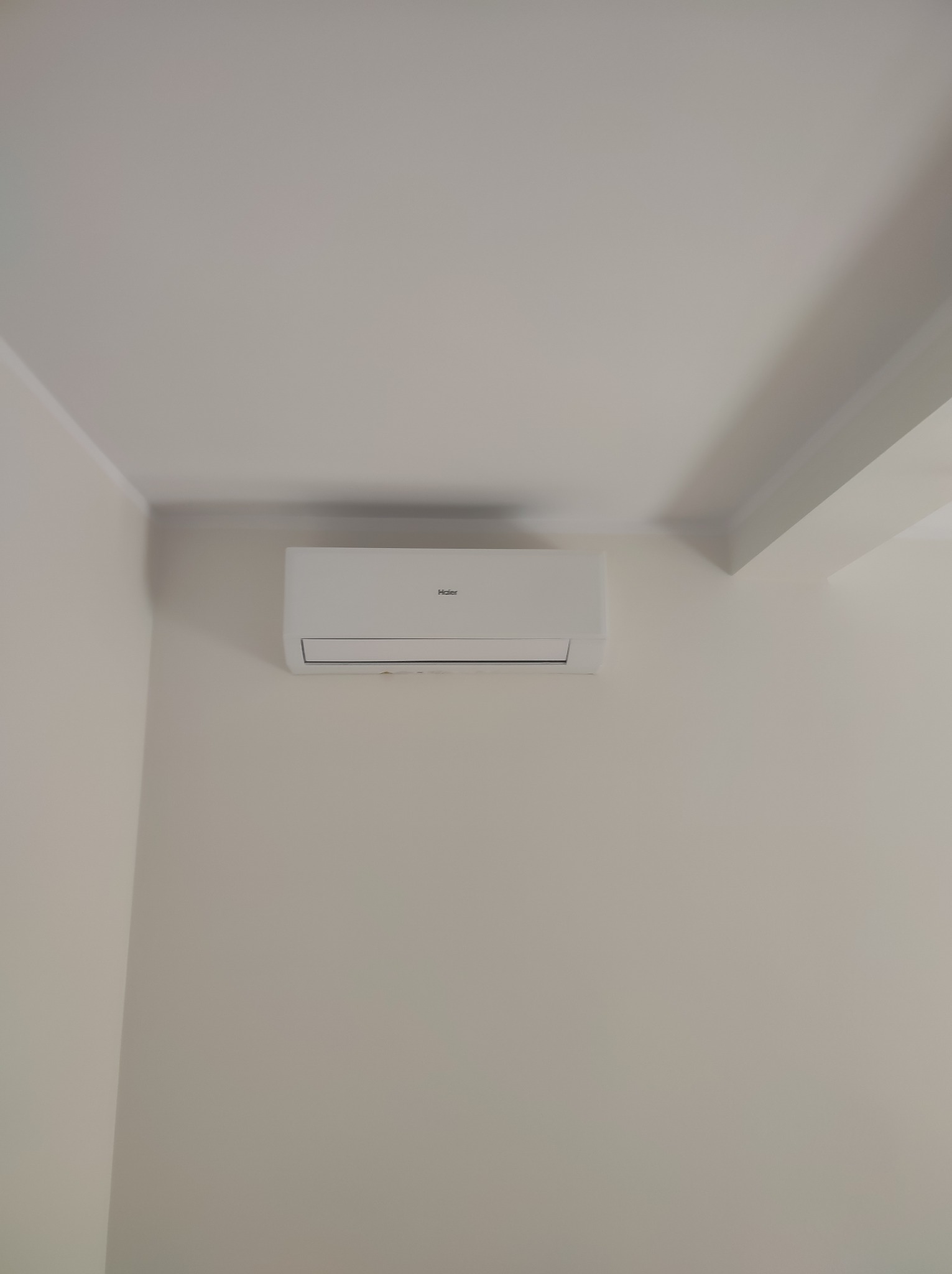 Biała jednostka klimatyzacji Haier zamontowana na kremowej ścianie w rogu pomieszczenia, widoczny fragment sufitu z dekoracyjną listwą.