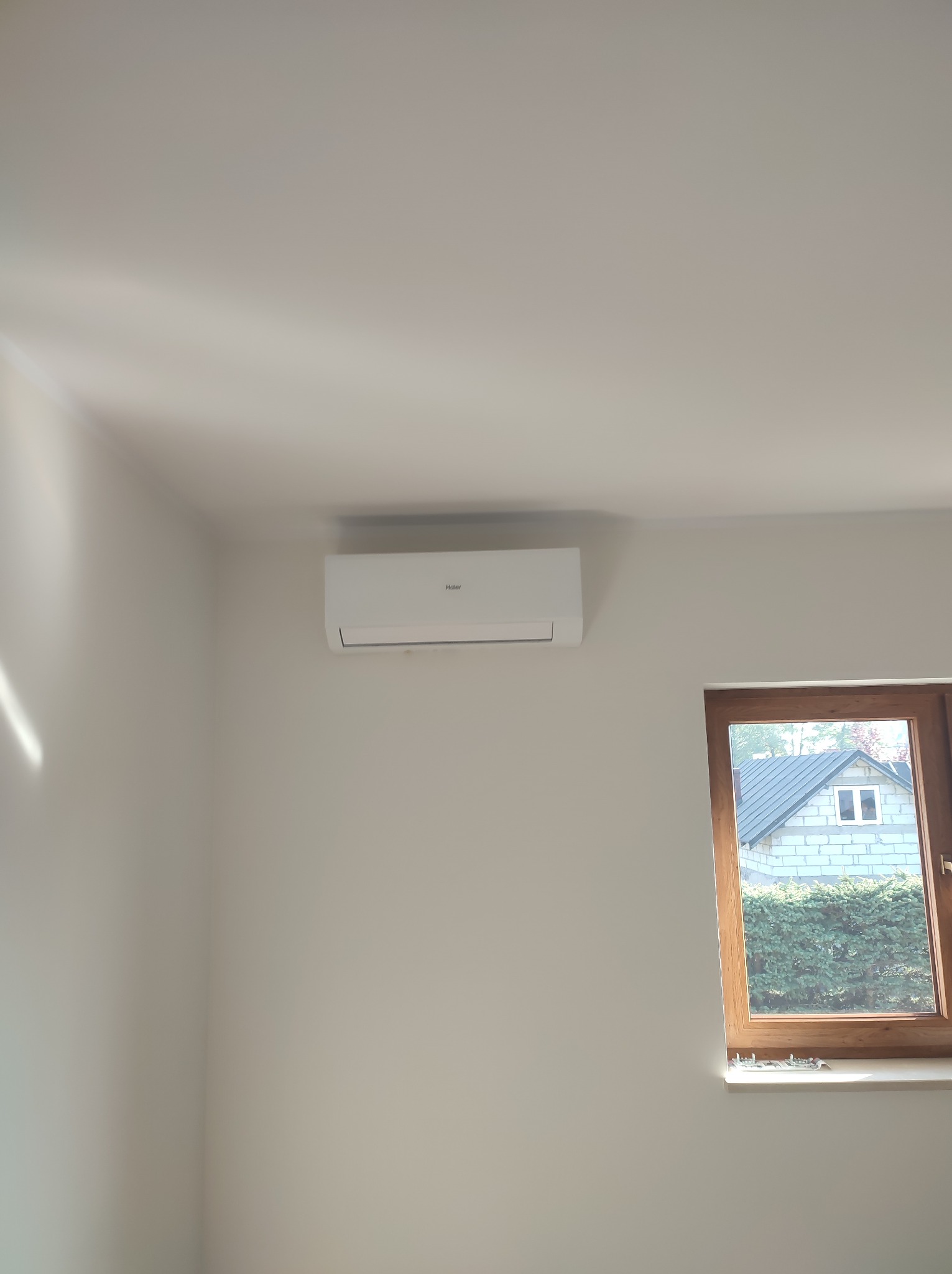 Biały klimatyzator Haier zamontowany na ścianie w jasnym pokoju z oknem, widok na budynek z szarej cegły i zielony żywopłot.
