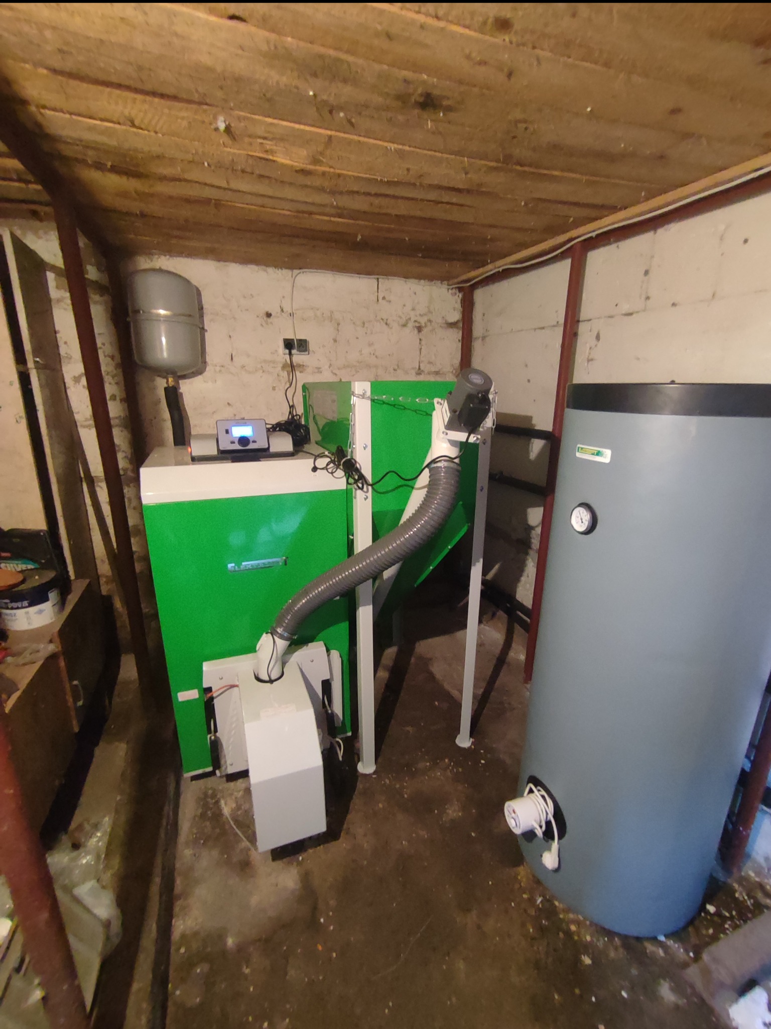 Zielony piec z podajnikiem i szary zbiornik akumulacyjny w kotłowni z drewnianym stropem i betonową podłogą.