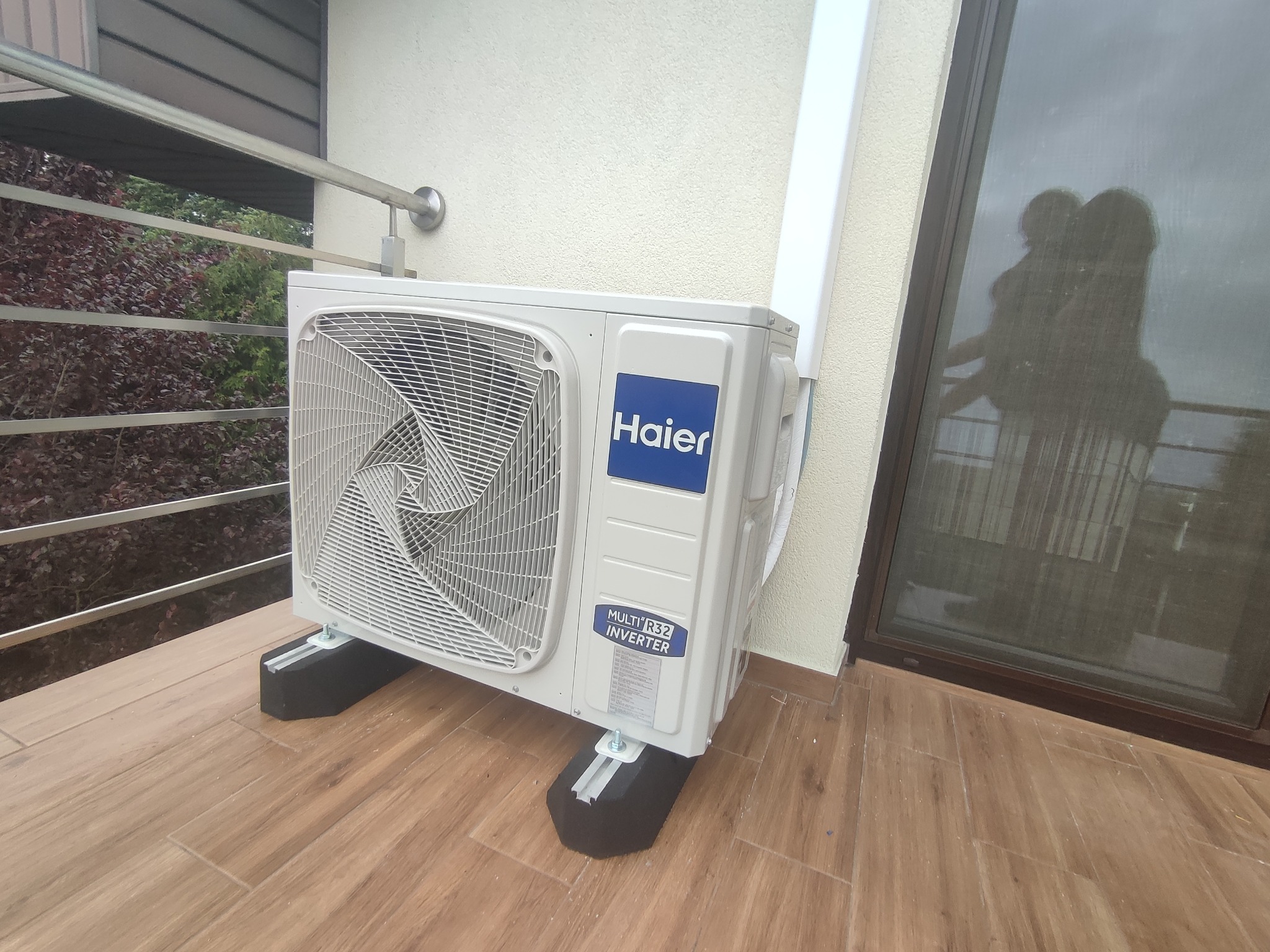 Zewnętrzna jednostka klimatyzacji Haier na balkonie z brązowymi płytkami, widoczny fragment balustrady i odbicie w oknie
