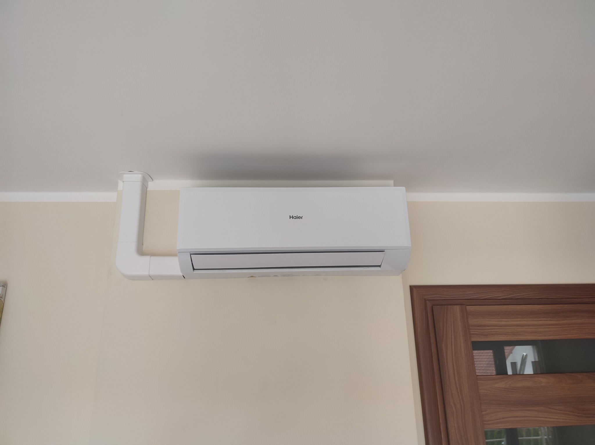 Biała klimatyzacja Haier zamontowana na beżowej ścianie, widoczny fragment brązowych drzwi i biała listwa przypodłogowa pod sufitem.