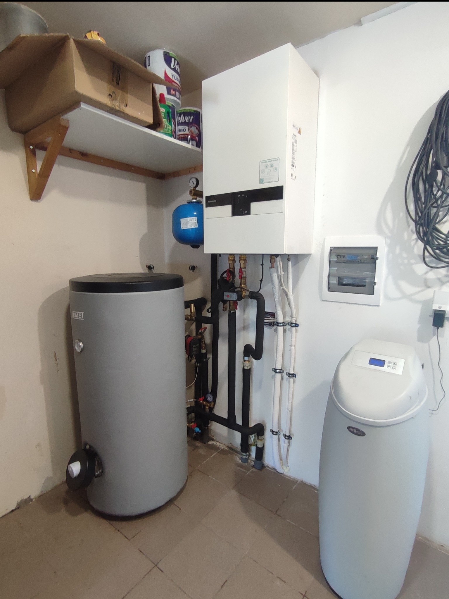 Kocioł grzewczy wiszący na ścianie obok szarego zasobnika wody i zmiękczacza wody, instalacja z rurami i zaworami, na półce karton i puszki z farbą.