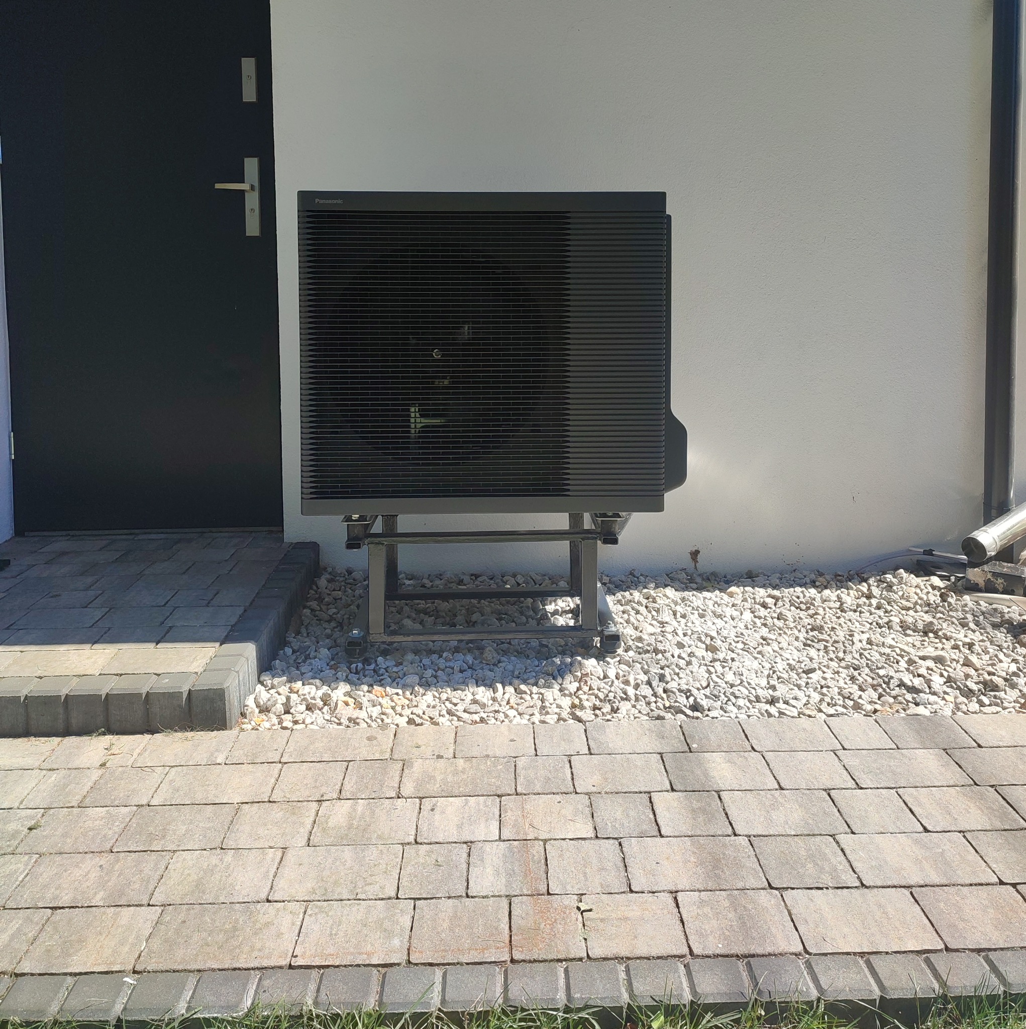 Czarna pompa ciepła Panasonic zamontowana na metalowej konstrukcji na zewnątrz budynku, obok ciemnych drzwi wejściowych i białej ściany, otoczona białymi kamieniami.