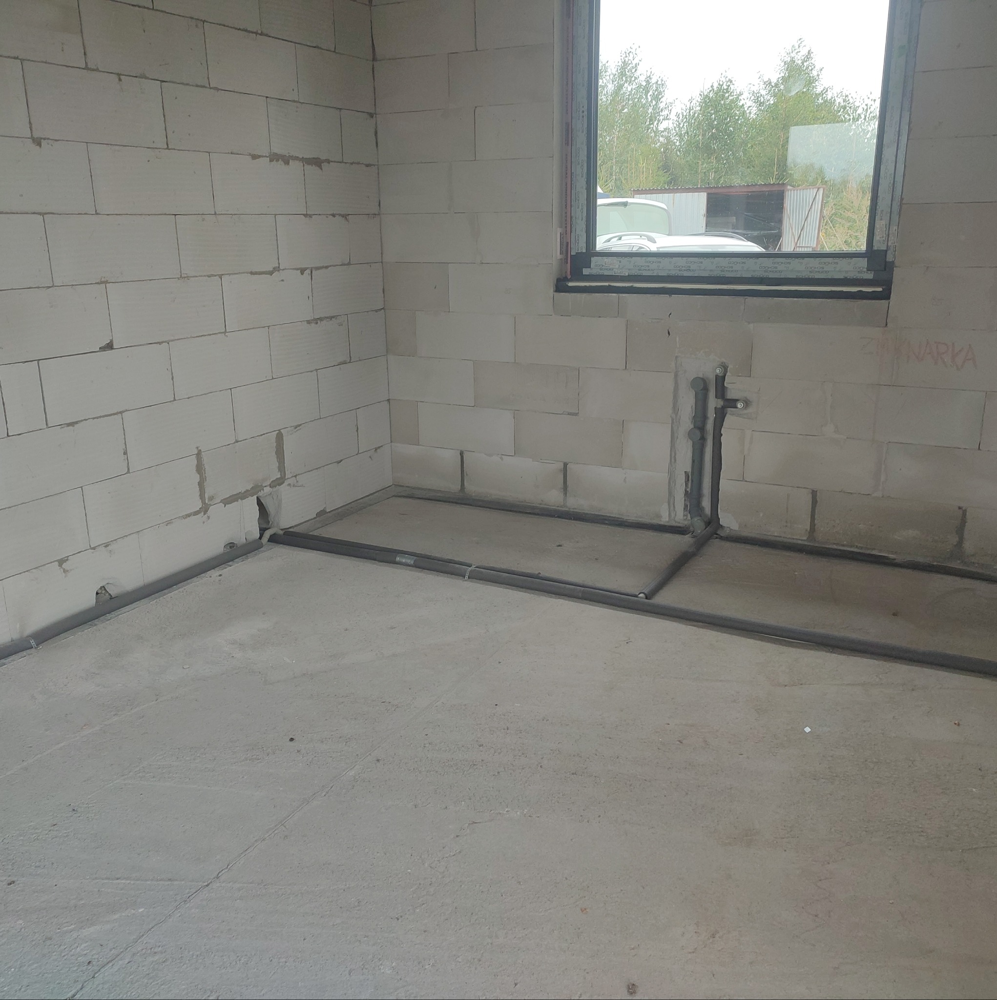 Surowy stan instalacji grzewczej w nowym budynku z widocznymi rurami prowadzonymi wzdłuż ścian i podłogi, okno z widokiem na zewnątrz.