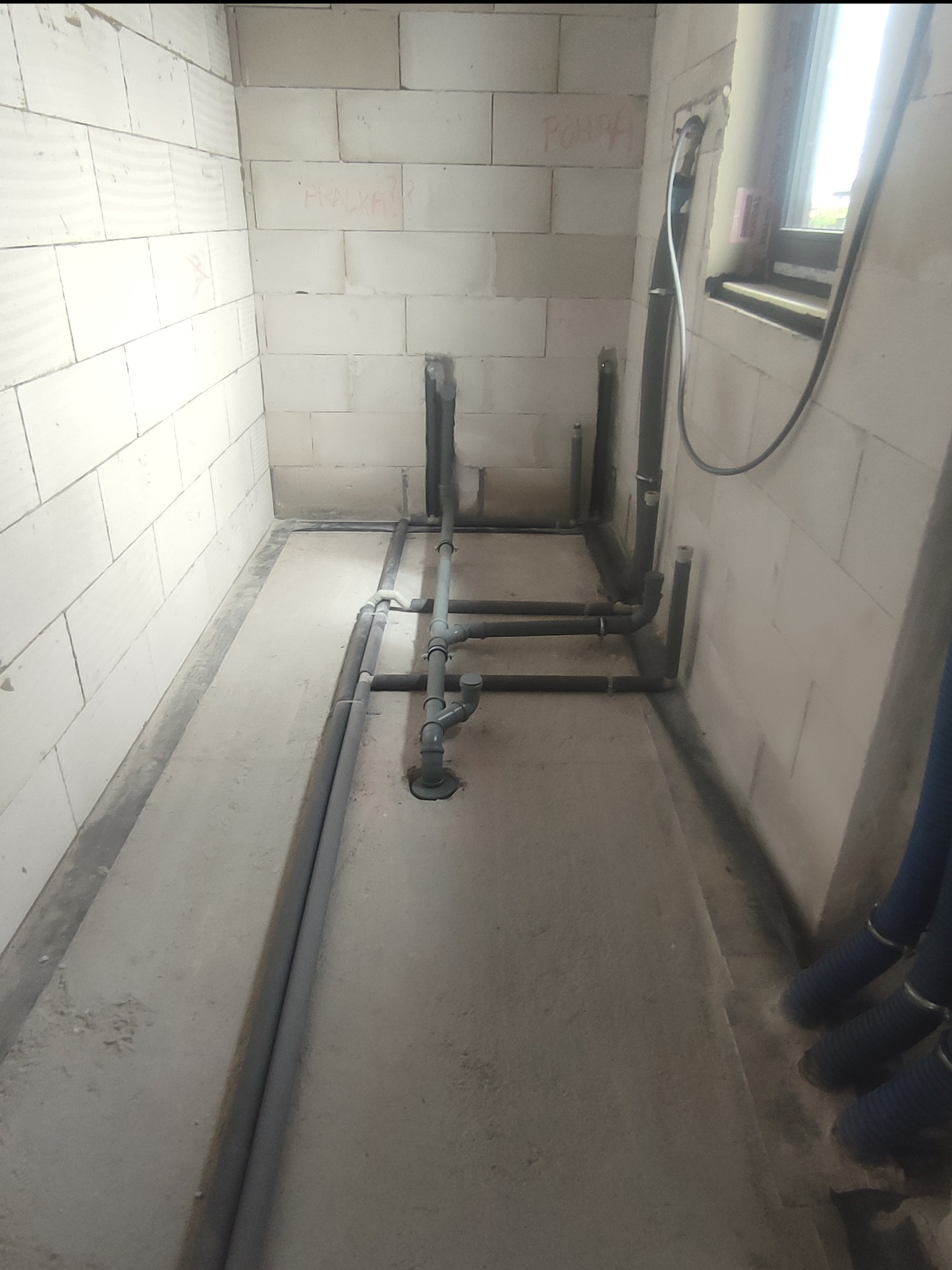 Szare rury instalacyjne ułożone na betonowej podłodze w surowym pomieszczeniu z białych bloczków, widoczne napisy na ścianie i fragment okna.