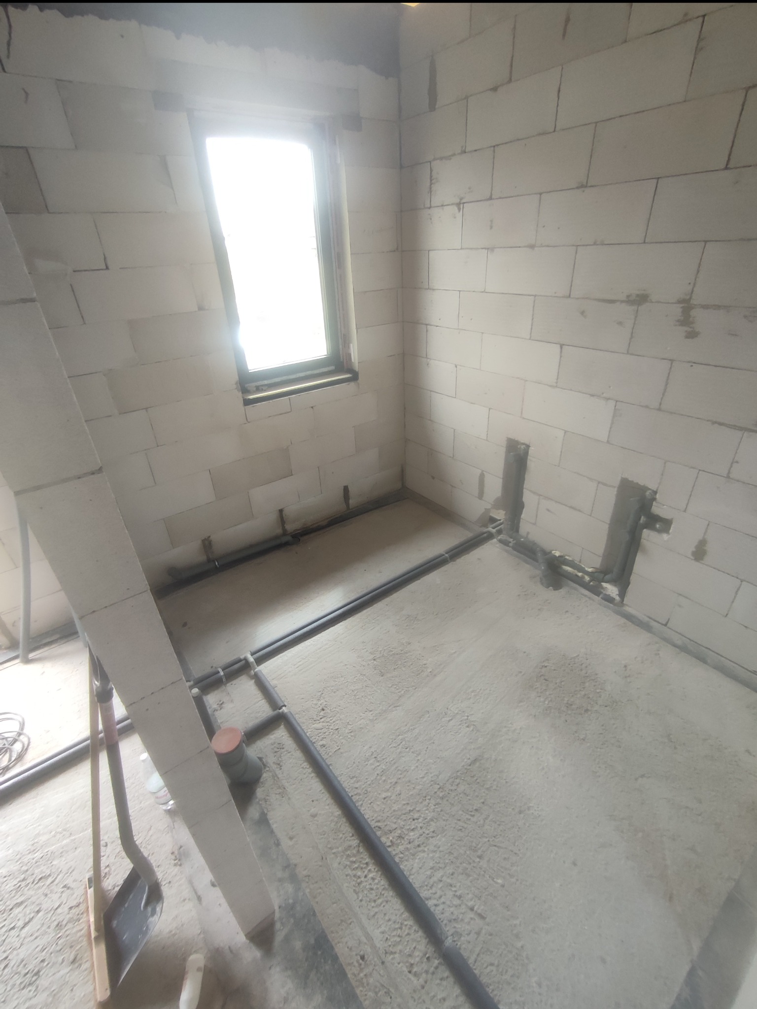 Surowy stan: czarne rury instalacji wodnej ułożone na posadzce w pomieszczeniu z białych bloczków, widoczne fragmenty narzędzi i okno.