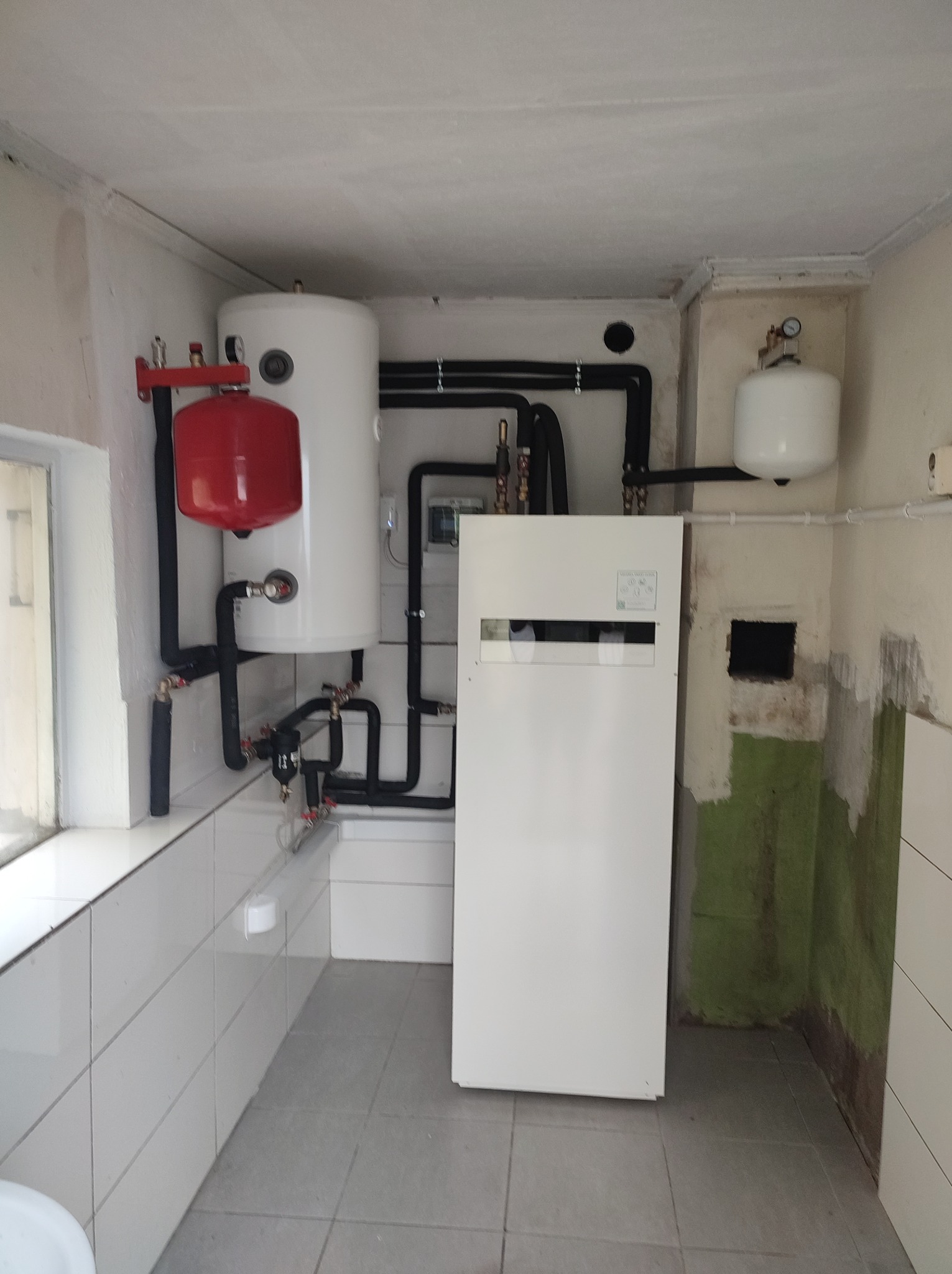 Instalacja grzewcza z białym zasobnikiem i czerwonym naczyniem wzbiorczym w pomieszczeniu w Toruniu, z widocznymi rurami i elementami mocującymi.