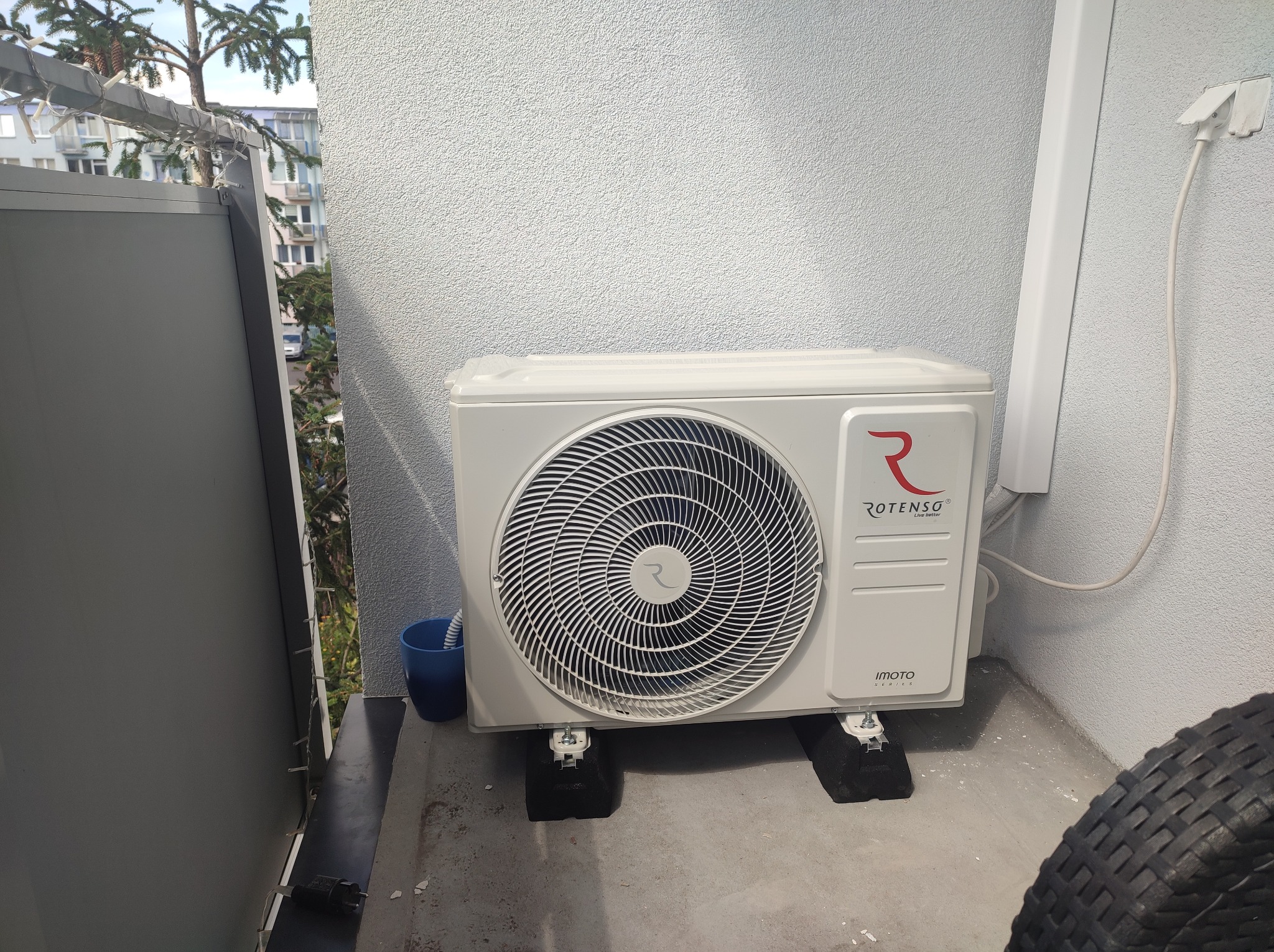 Biała jednostka zewnętrzna klimatyzacji Rotenso Imoto Series zamontowana na czarnych podkładkach antywibracyjnych na balkonie, widoczny fragment opony i niebieski kubek.
