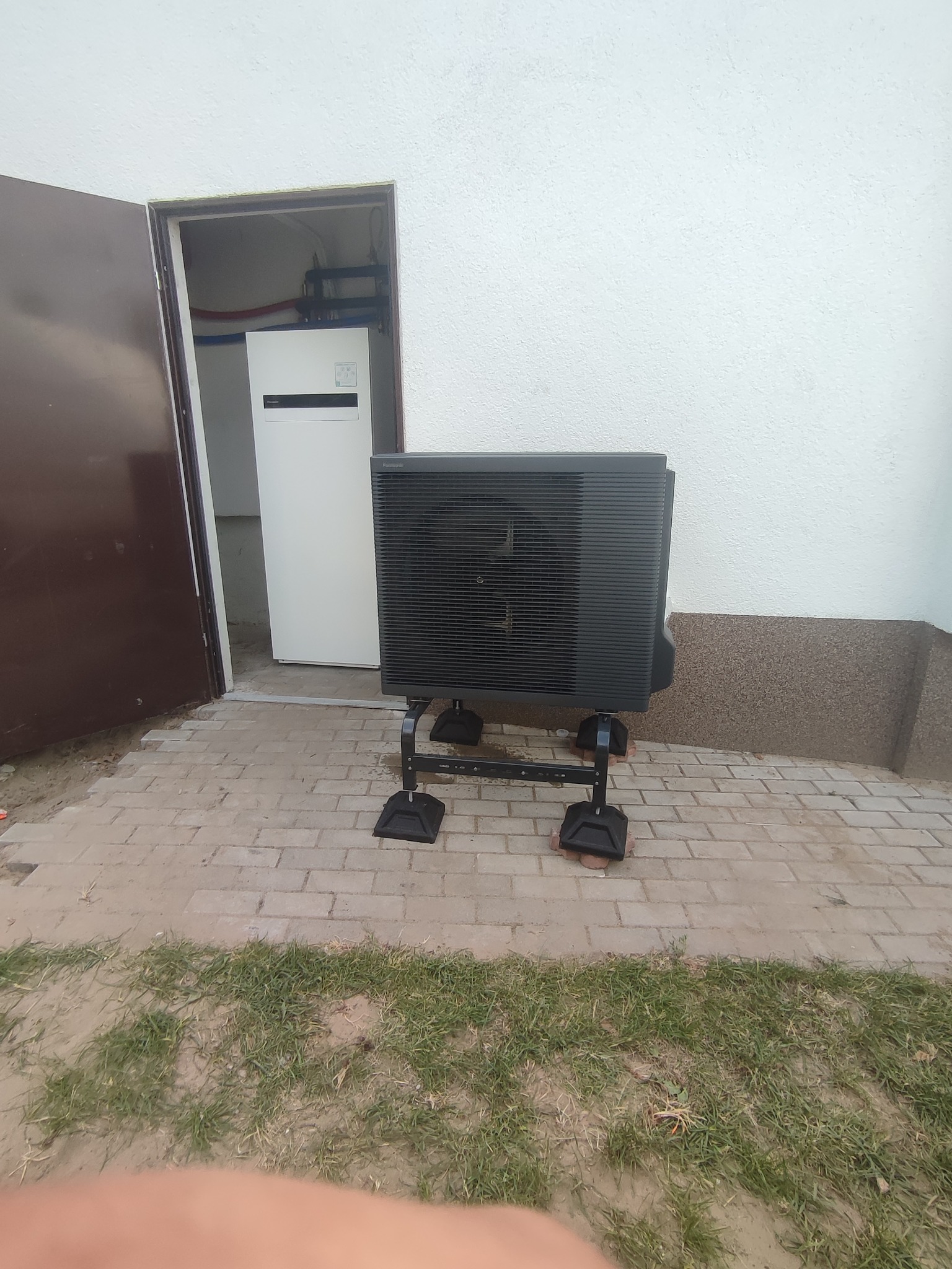 Czarna zewnętrzna jednostka pompy ciepła Panasonic Aquarea na metalowym stelażu, ustawiona na kostce brukowej przed białą ścianą budynku, z otwartymi drzwiami do pomieszczenia z wewnętrzną jednostką.