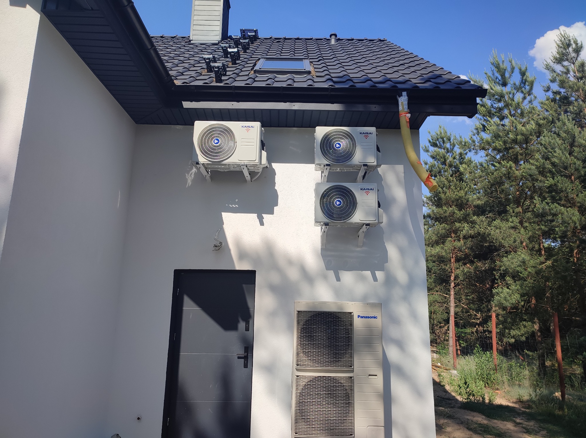 Zewnętrzne jednostki klimatyzacji Kaisai i Panasonic zamontowane na białej ścianie budynku z ciemnoszarą stolarką okienną i dachem, widoczne przewody odprowadzające skropliny.