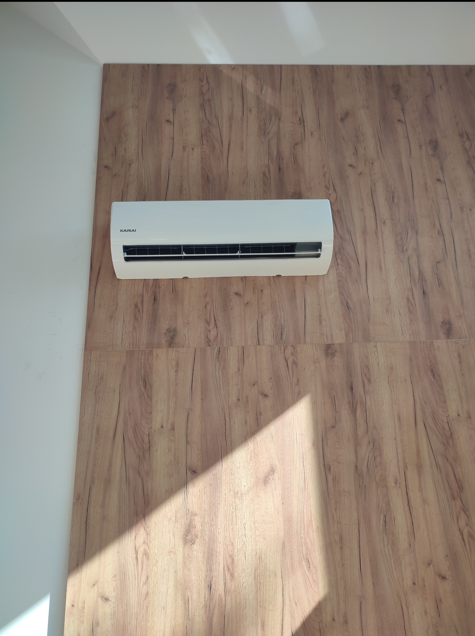 Biały klimatyzator marki Kaisai zamontowany na ścianie wyłożonej panelami imitującymi drewno. Widoczny cień padający na panele.