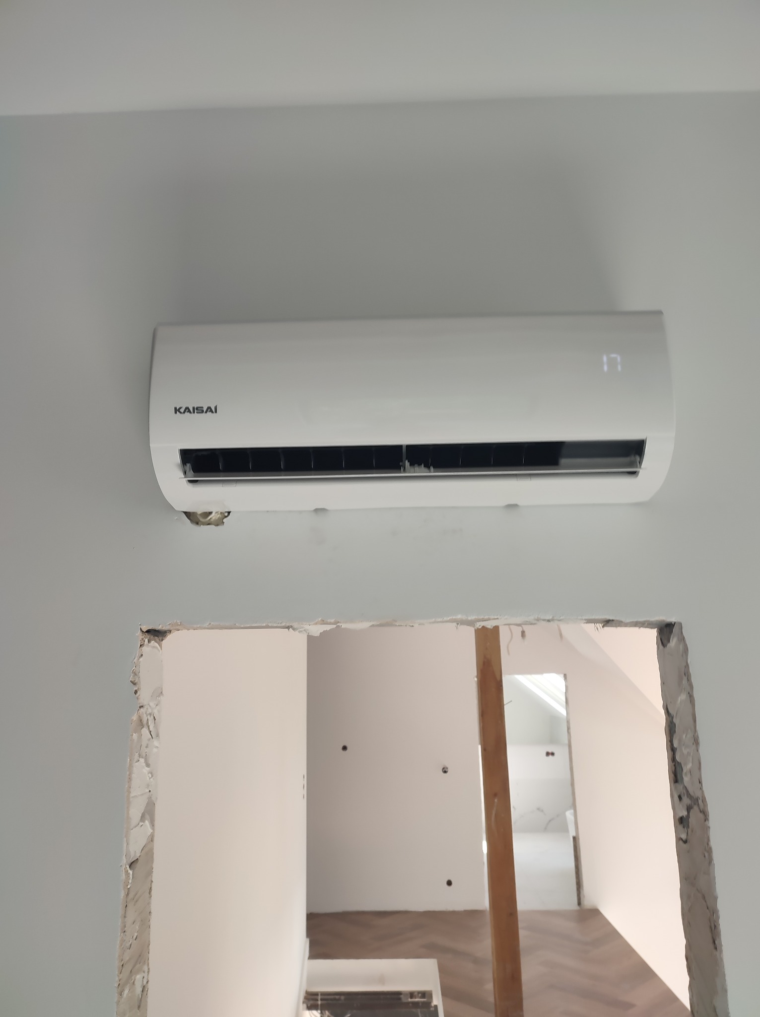 Biały klimatyzator Kaisai zamontowany na szarej ścianie, widoczne niedokończone prace wykończeniowe wokół otworu w ścianie.