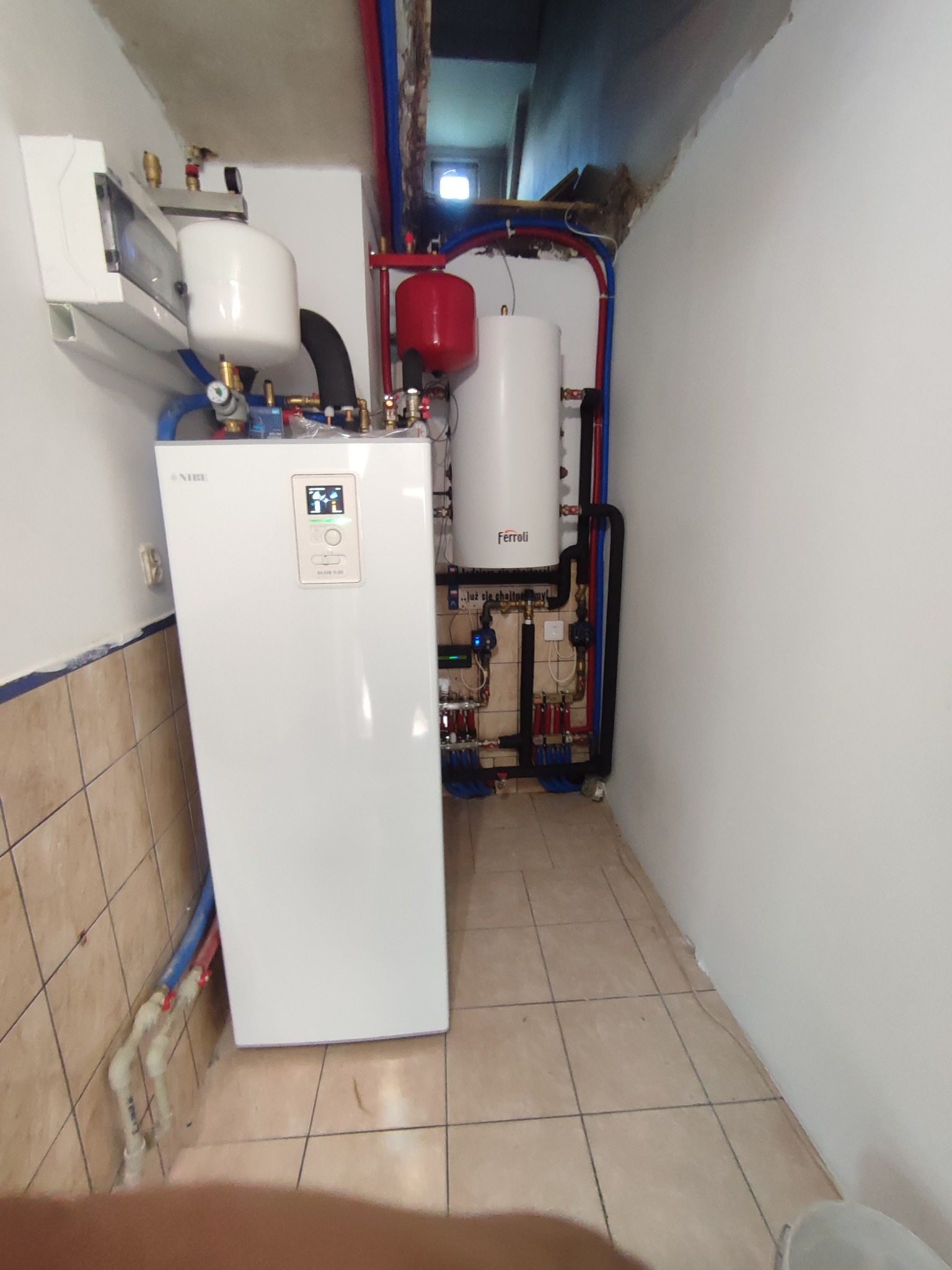 Biała pompa ciepła Nibe z podłączonymi rurami instalacji grzewczej, obok zbiornik Ferroli, widoczne elementy instalacji w ciasnym pomieszczeniu.