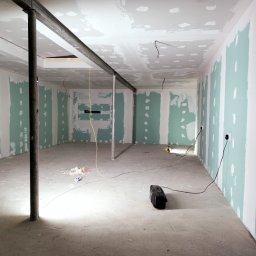 Damian building service - Surowe wnętrze w trakcie remontu: ściany pokryte płytami karton gips z widocznymi łączeniami do szpachlowania, metalowe podpory stropu, rozproszone kable elektryczne i narzędzia, betonowa podłoga...