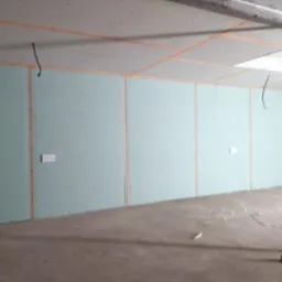 Wnętrze w trakcie prac wykończeniowych: surowe ściany z płyt kartonowo-gipsowych, widoczne łączenia i gniazdka elektryczne, betonowa podłoga z resztkami materiałów budowlanych, instalacja pod sufitem.