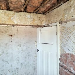 Damian building service - Pokój w trakcie remontu: odsłonięta izolacja stropu, fragmenty starej farby na ścianach, białe drzwi z kablem zwisającym nad górną krawędzią.