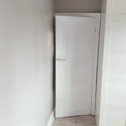 Damian building service - Wnętrze po remoncie: białe, częściowo otwarte drzwi z poziomymi panelami, srebrna klamka, szara ściana pomalowana farbą w odcieniach szarości, podłoga z paneli imitujących jasne drewno, biała szafa...