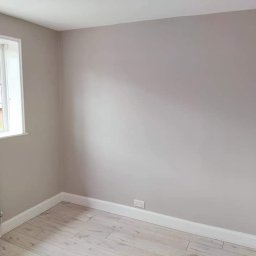 Damian building service - Pomalowane na szaro wnętrze pokoju z jasnymi panelami podłogowymi, białymi listwami przypodłogowymi i grzejnikiem pod oknem.