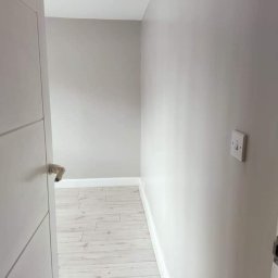 Damian building service - Korytarz po odświeżeniu, ściany pomalowane na jasny odcień szarości, białe listwy przypodłogowe, panele podłogowe imitujące bielone drewno, widoczny włącznik światła.