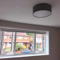 Damian building service - Świeżo pomalowane wnętrze pokoju z białym oknem PCV, częściowo otwartym, widok na ulicę z ceglanymi domami i samochodami, szary abażur lampy sufitowej.