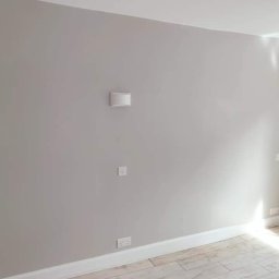 Damian building service - Świeżo pomalowane wnętrze pokoju w odcieniach szarości, widoczne gniazdka elektryczne i lampa ścienna, wykończone listwy przypodłogowe i panele podłogowe imitujące drewno.
