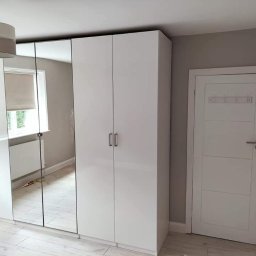 Damian building service - Biała, lakierowana szafa wnękowa z lustrzanymi drzwiami, zamontowana w pokoju z drewnianą podłogą i białą, panelową ścianą, widoczna lampa sufitowa.