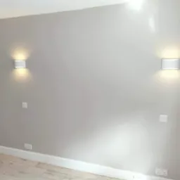 Jasnoszara ściana z dwoma nowoczesnymi, białymi kinkietami, widoczne gniazdka elektryczne i listwy przypodłogowe z jasnego drewna.