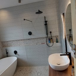 Damian building service - Wykończona łazienka w Dwikozach z wanną wolnostojącą, umywalką nablatową na drewnianym blacie, czarną armaturą i mozaiką na ścianach i podłodze. Widoczne okno z widokiem na zieleń.