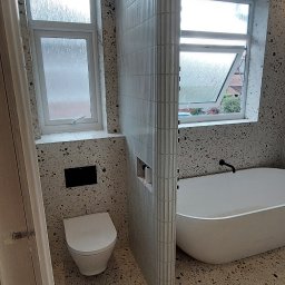 Damian building service - Wykończona łazienka w stylu lastryko z białą wolnostojącą wanną, toaletą podwieszaną i ścianką działową z pionowych płytek w odcieniach szarości.