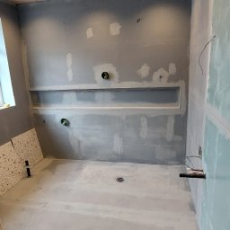 Damian building service - Łazienka w trakcie remontu: przygotowane ściany z wnękami pod zabudowę, surowy beton na podłodze, widoczny odpływ liniowy, fragment płytek na podłodze i wiadro niebieskiej farby.