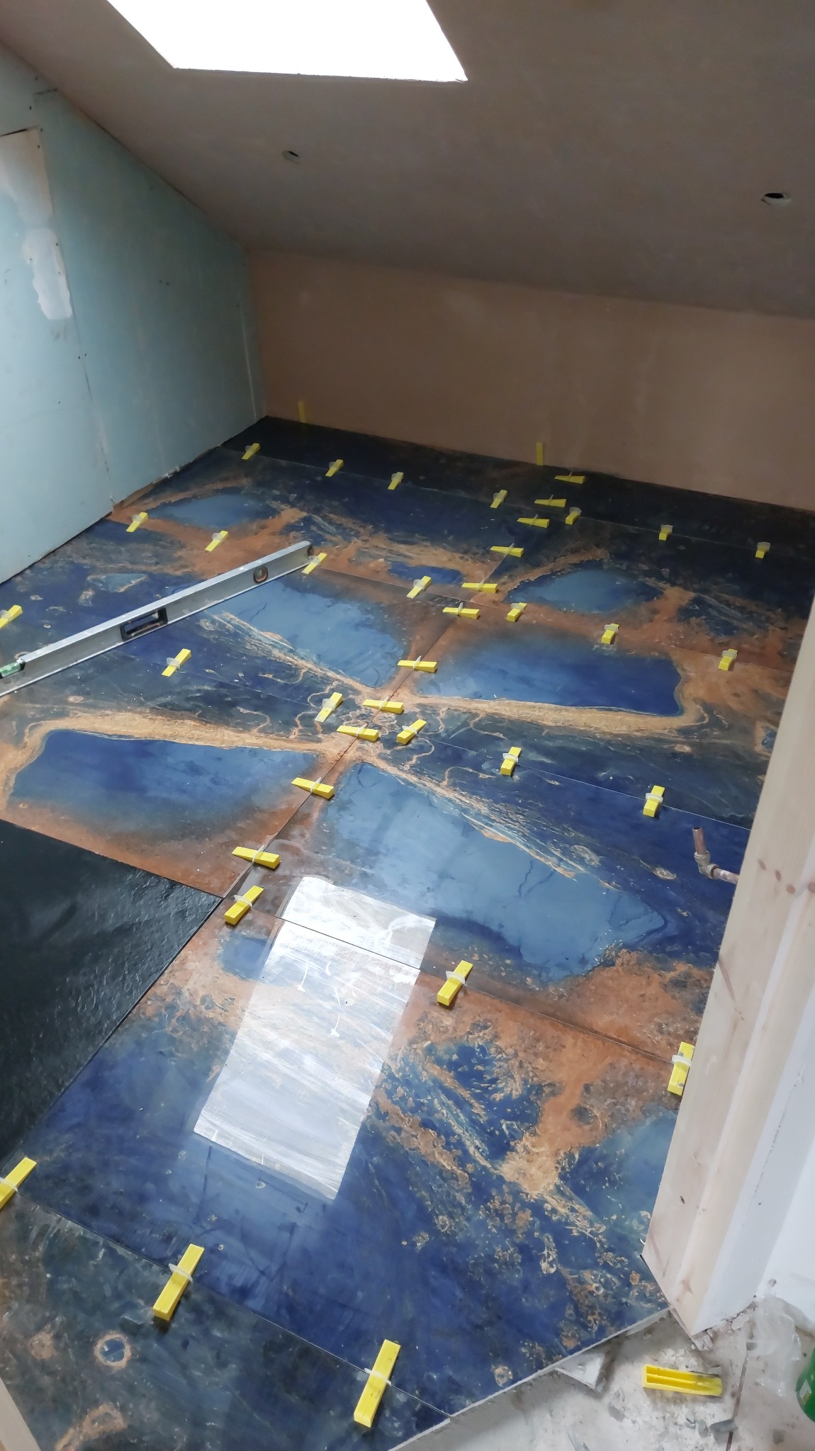 Układanie płytek w łazience na poddaszu z użyciem poziomicy i klipsów poziomujących; widoczne niebiesko-brązowe płytki imitujące marmur.