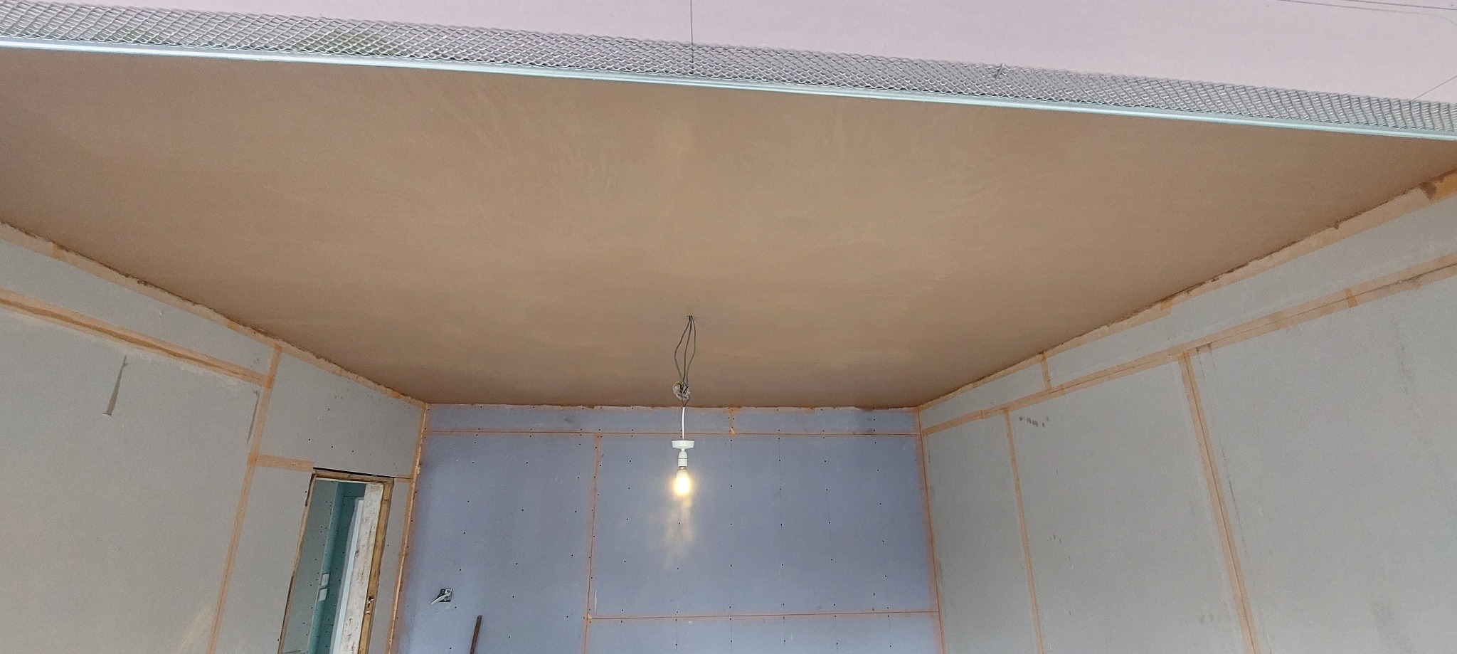Świeżo wyszpachlowany sufit i ściany z płyt kartonowo-gipsowych w trakcie remontu pomieszczenia, widoczne metalowe profile i prowizoryczne oświetlenie żarówką.